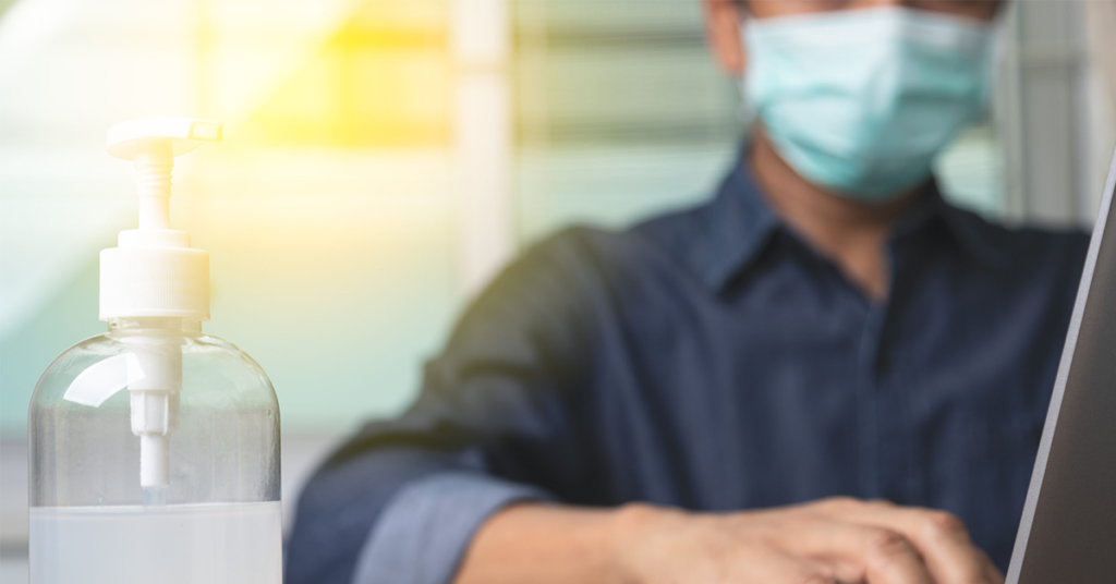 man working on laptop wearing a mask with bottle of hand sanitizer on desk